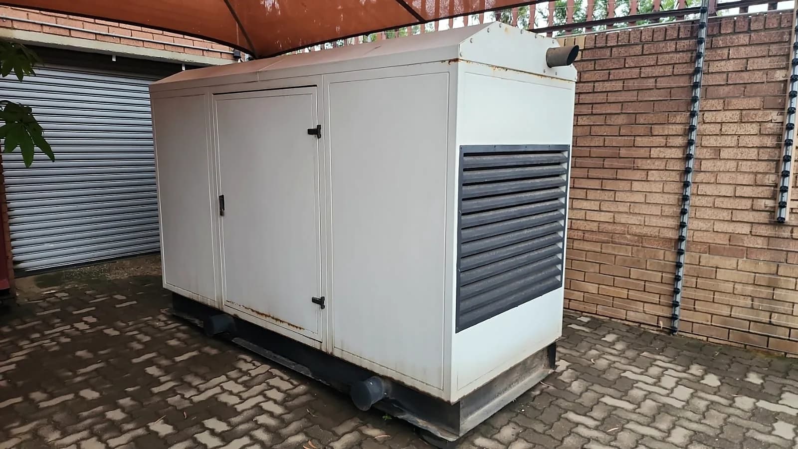 White canopied diesel generator under a shade-net carport next to a roller door