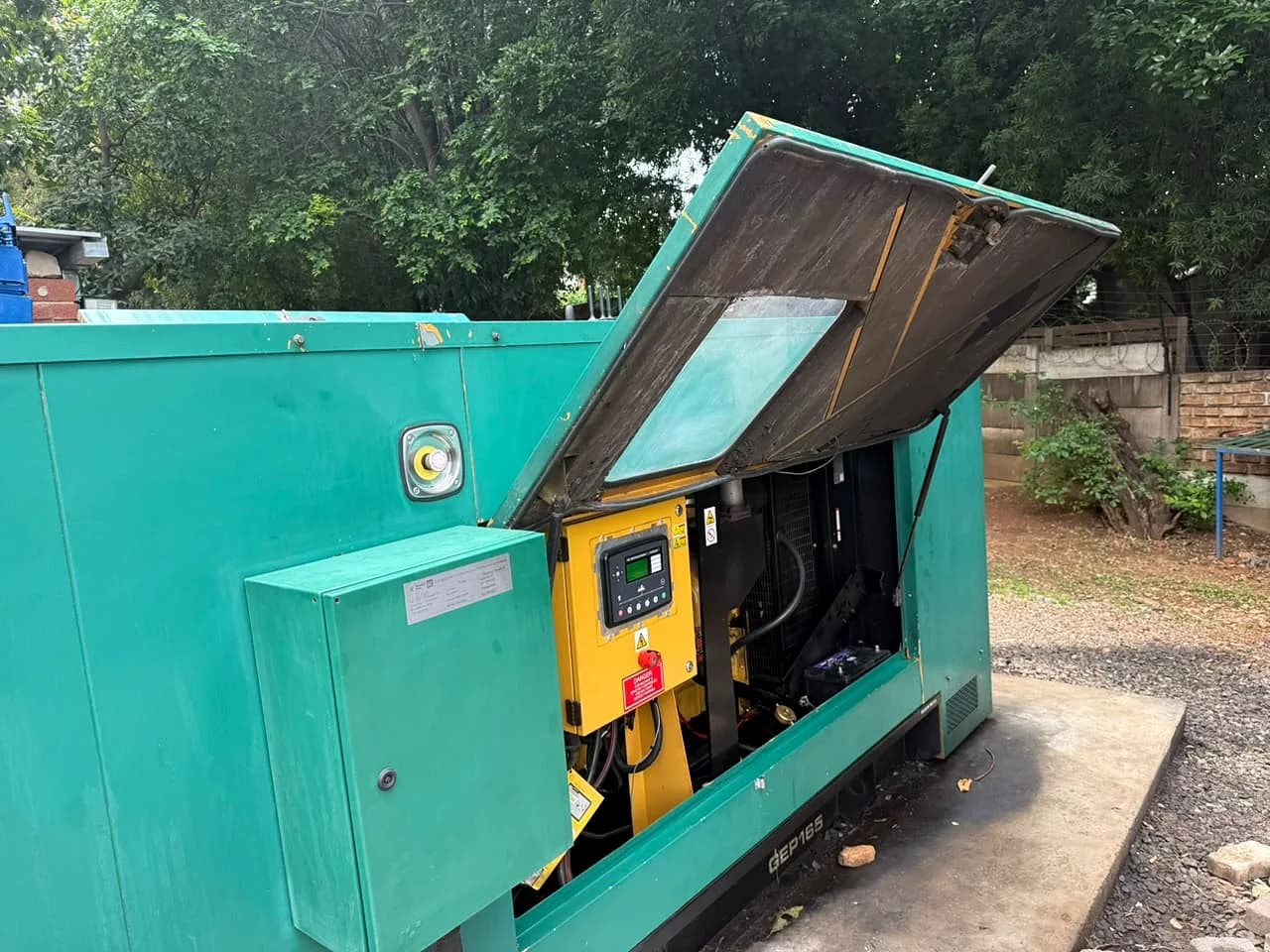 Teal diesel generator with its canopy lid raised, showing the engine control module and emergency stop in a yellow control box