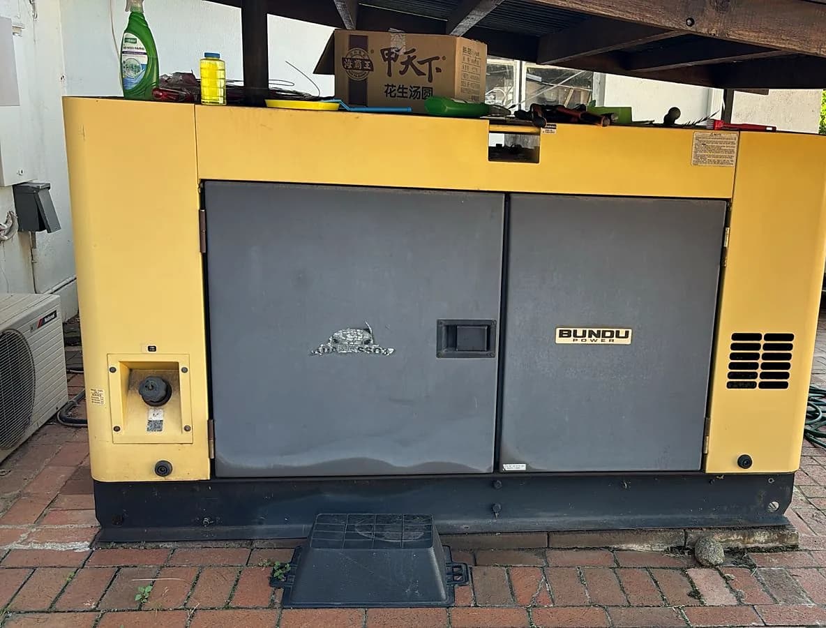 Yellow and grey Bundu Power silent generator under a carport, with bottles and boxes left on its roof
