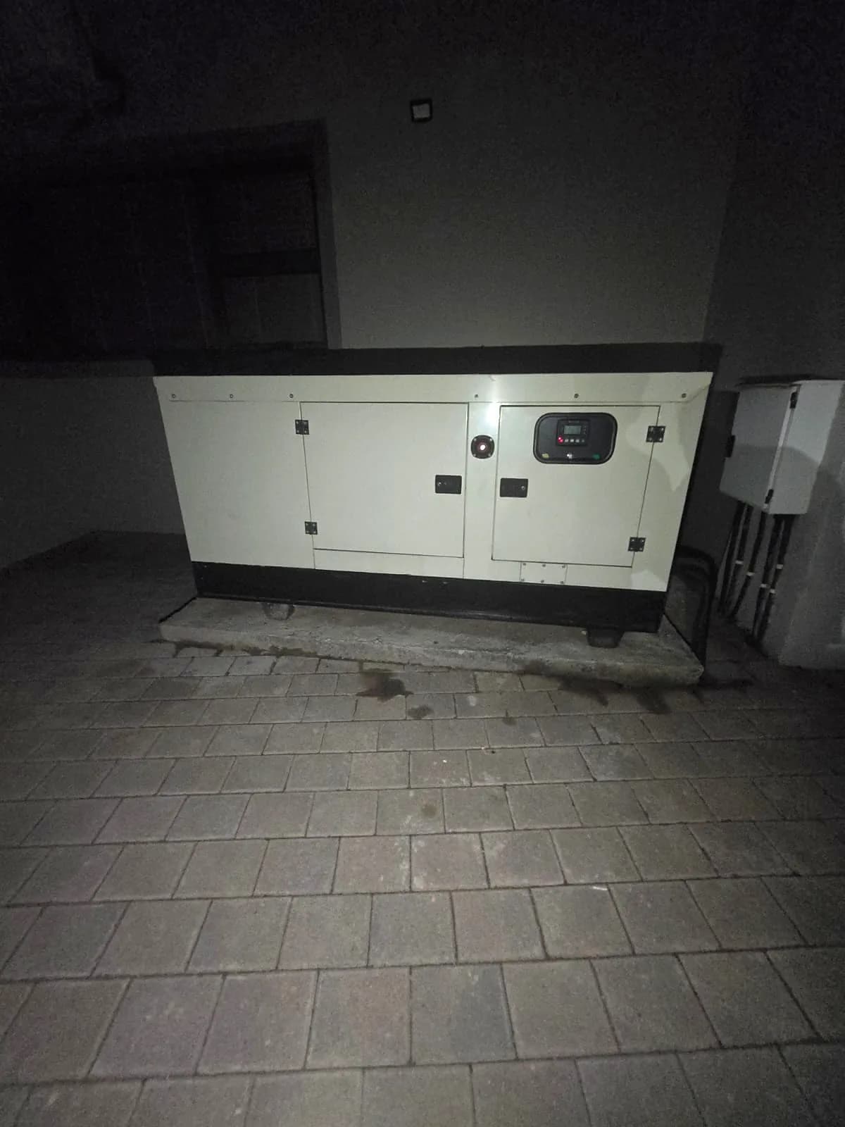 Diesel standby generator in a paved yard at night, lit by a torch, with oil marks on the paving in front of it