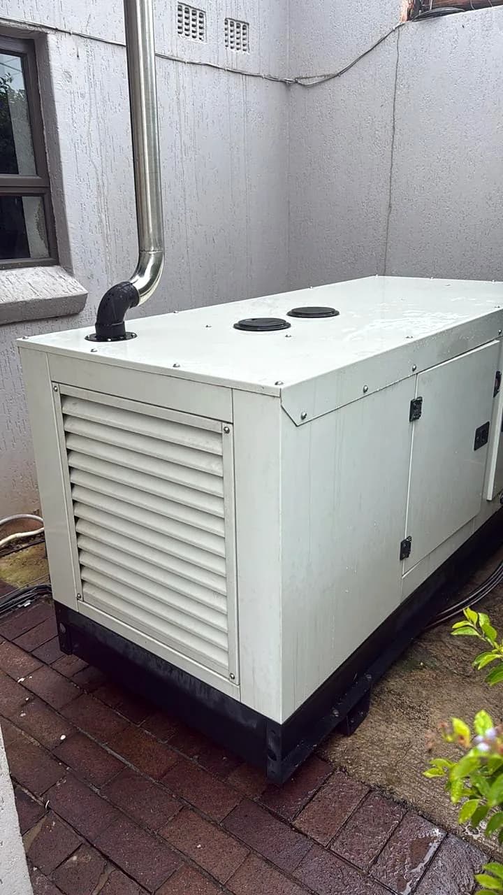 White silent generator in a narrow side passage of a house, with its exhaust pipe routed up the wall
