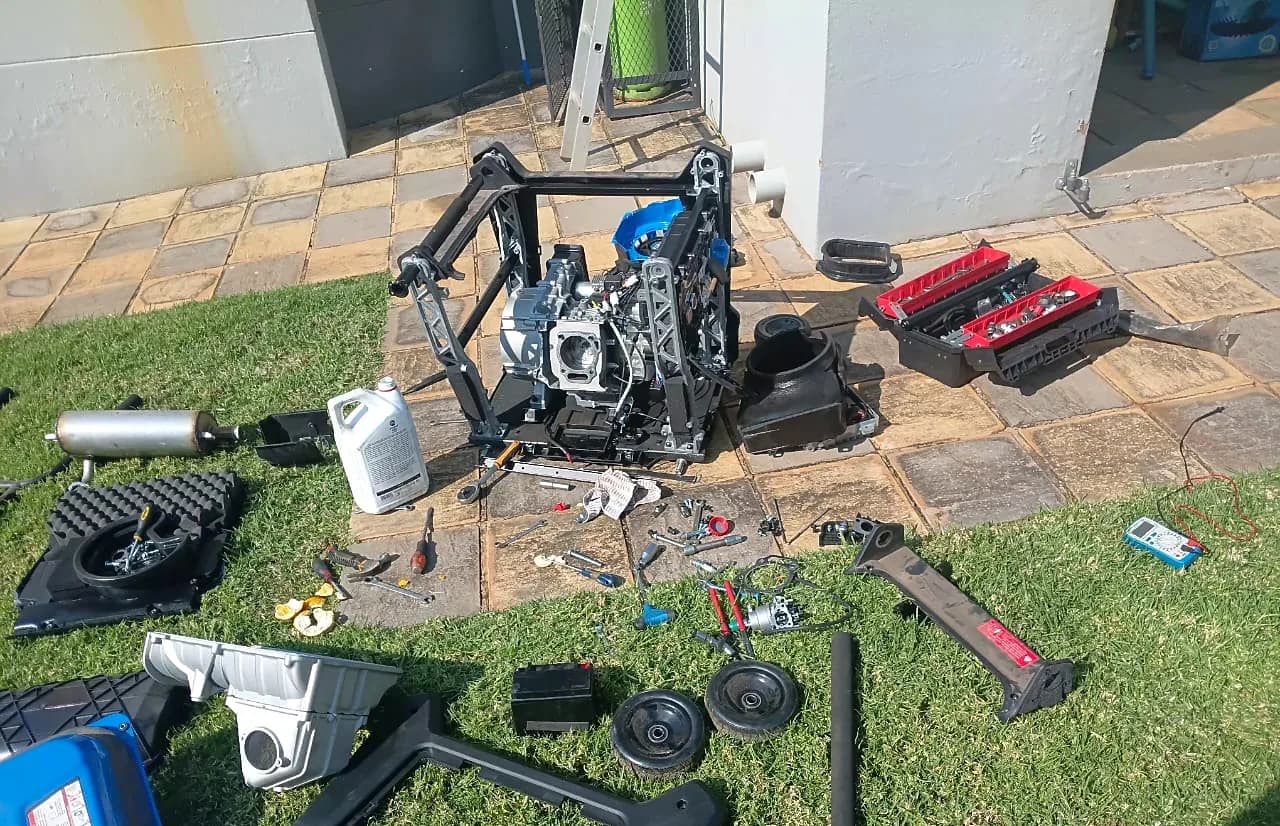 Portable petrol generator stripped down on a lawn for repair, with the engine, frame, fuel tank, parts, toolbox and multimeter laid out